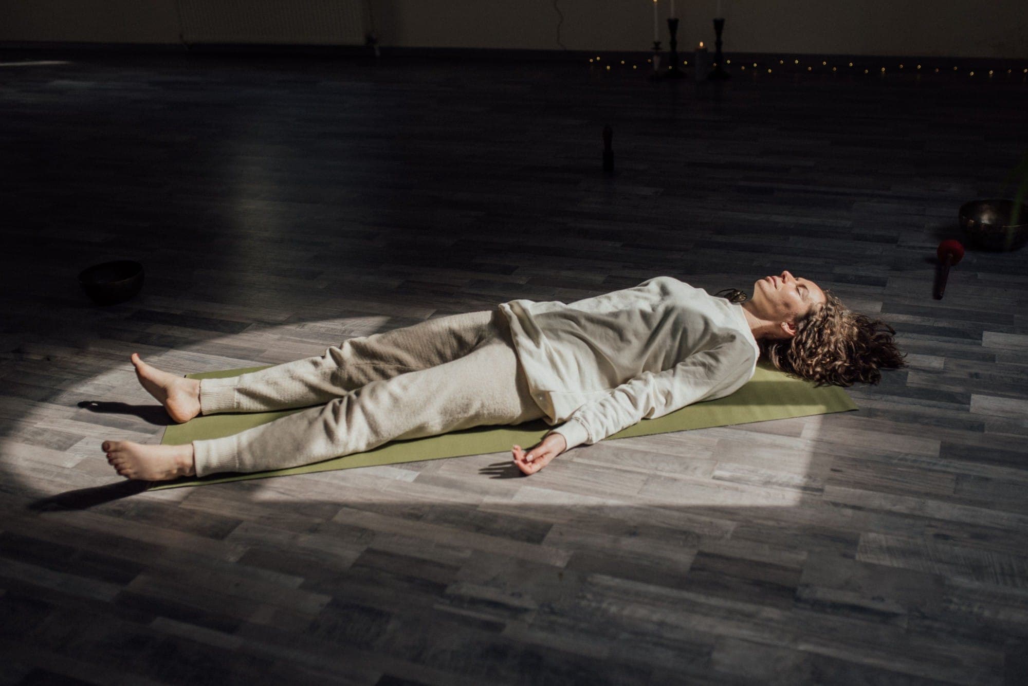 A woman lying at rest on a yoga mat in a pool of sunlight, singing bowls on the floor nearby
