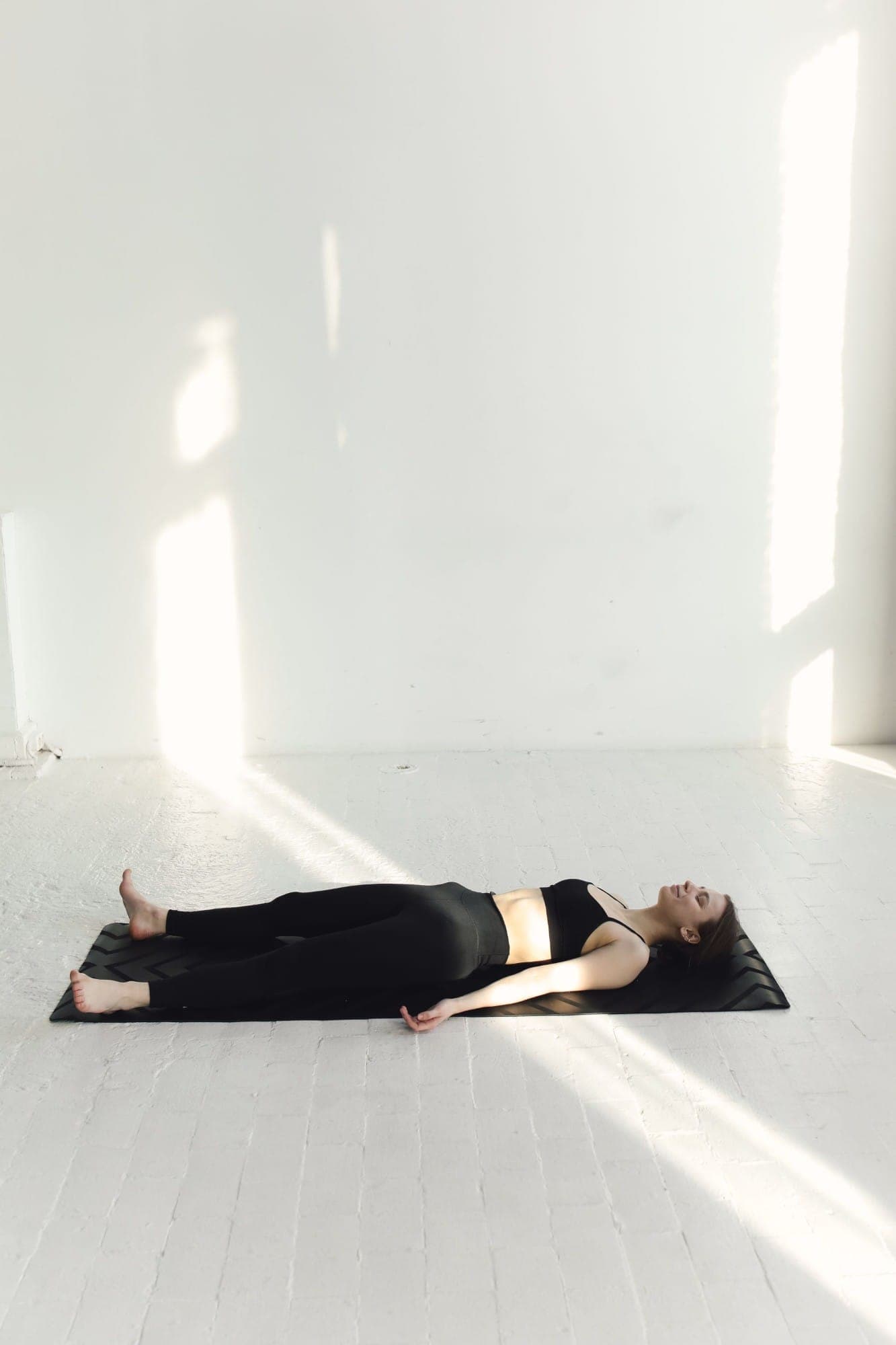 A woman lying at rest on a mat in a bright white room, sunlight across the floor