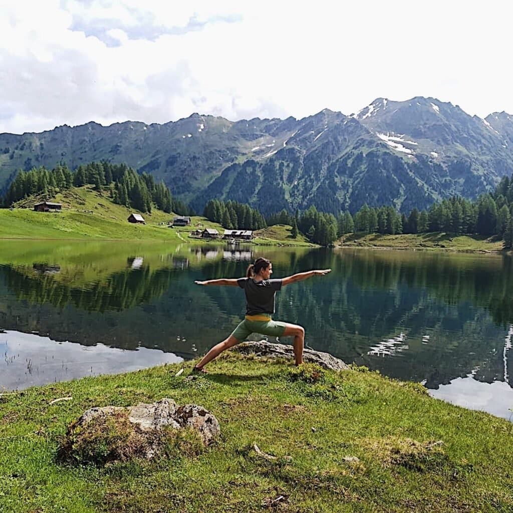 A woman in warrior pose on a grassy ridge above an alpine lake