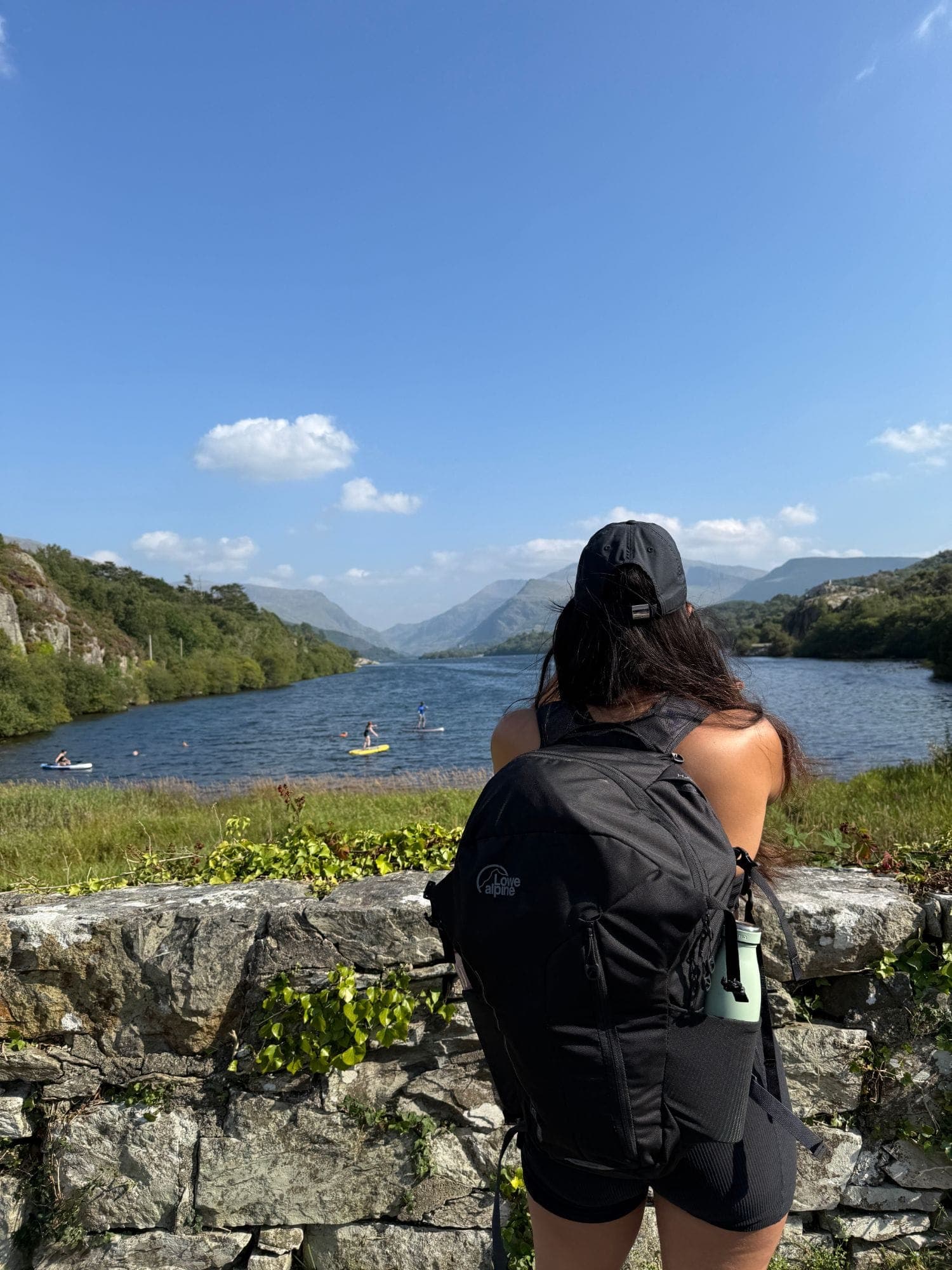 A walker with a rucksack looks out over the lake and mountains from a stone wall