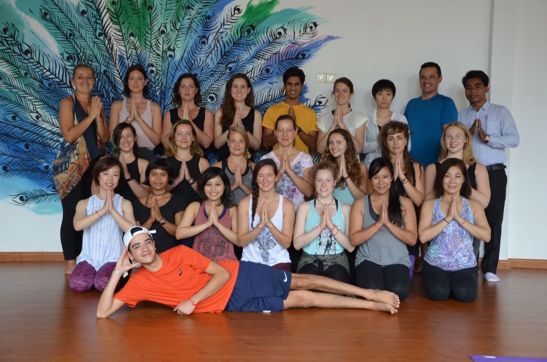 A teacher training group in namaste with Yogi Madhav, in front of a peacock mural