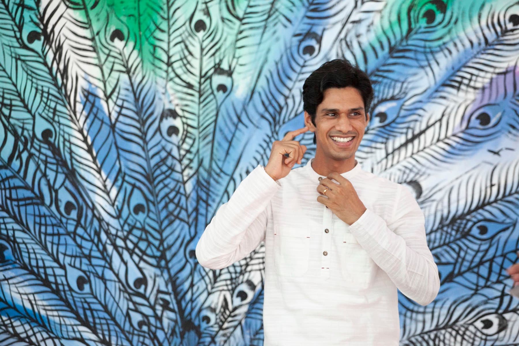 Yogi Madhav laughing in a white kurta in front of a painted peacock mural