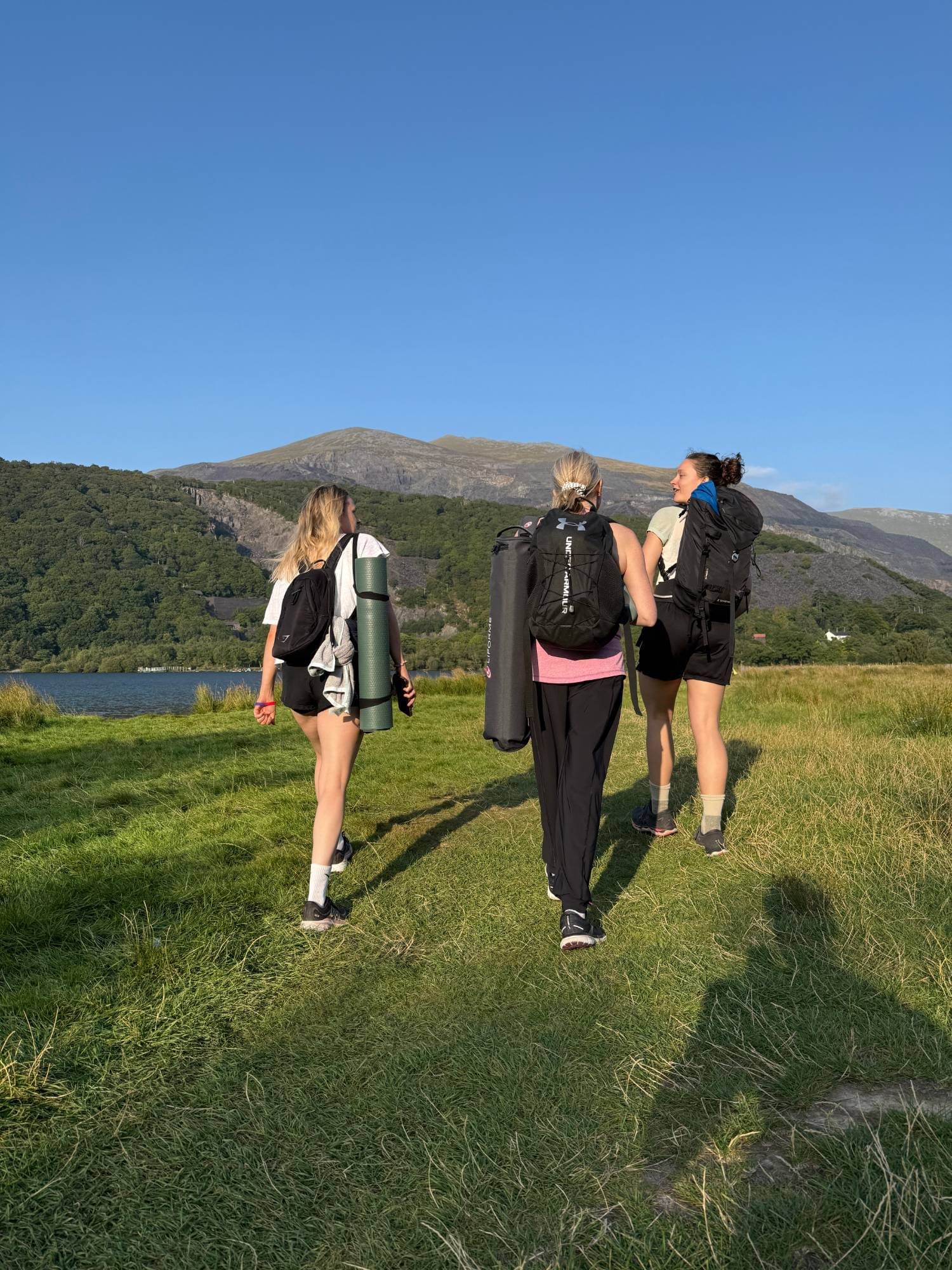Three people carrying yoga mats walk across a lakeside meadow towards the mountains