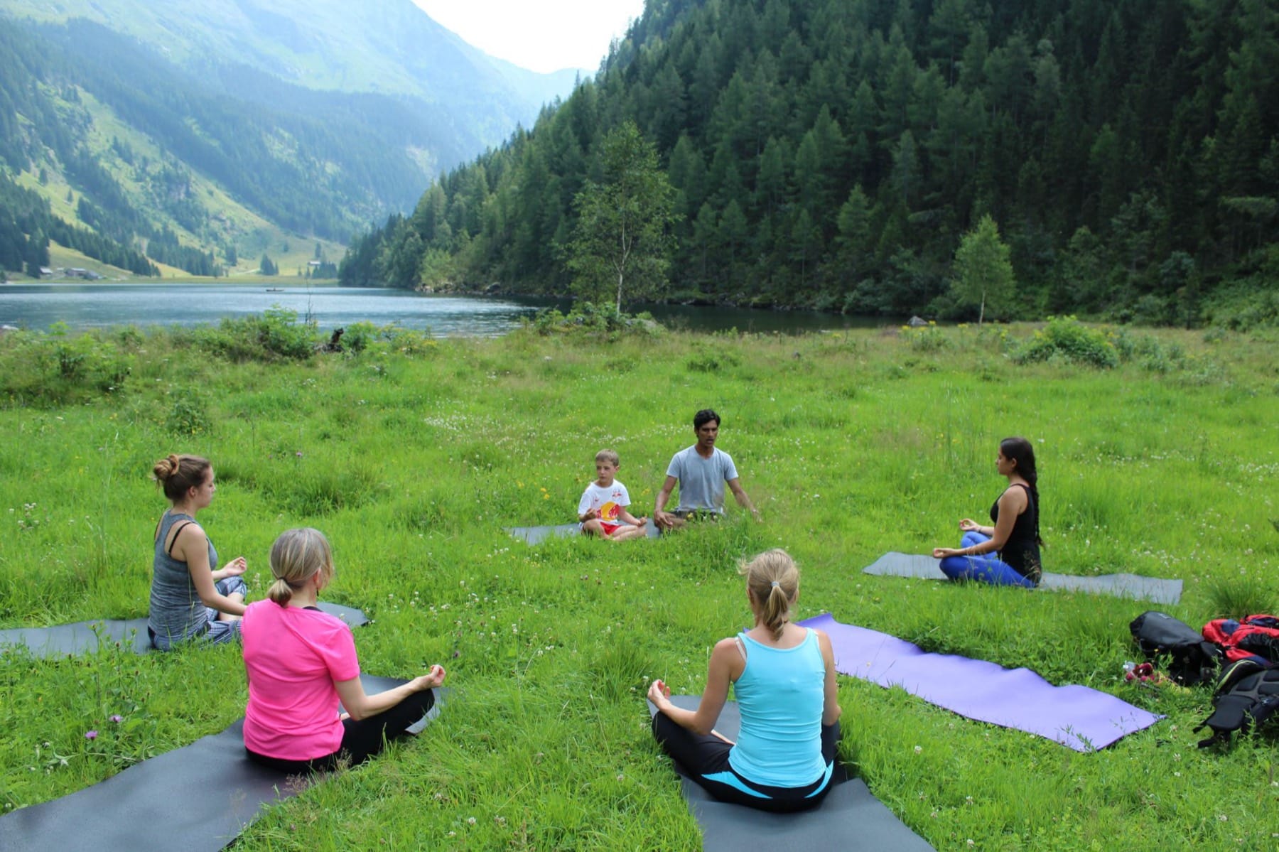 Yogi Madhav leading a seated meditation circle on a meadow beside an alpine lake in Austria