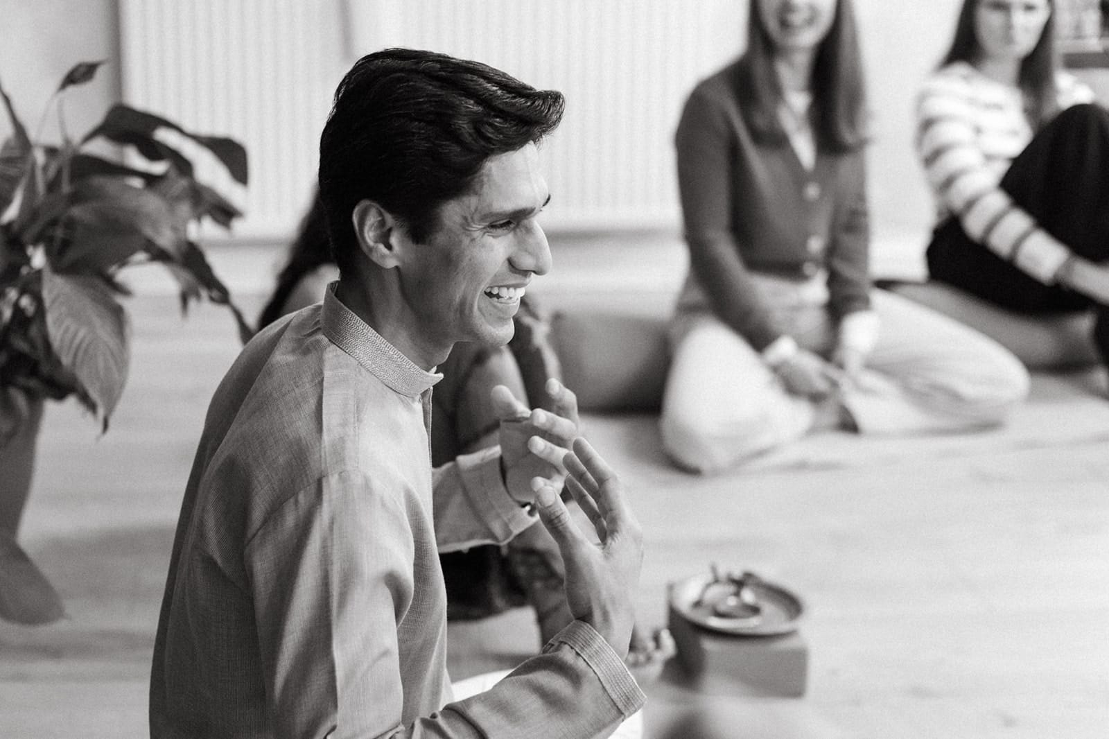 Yogi Madhav laughing as he explains something to students seated on the studio floor, in black and white