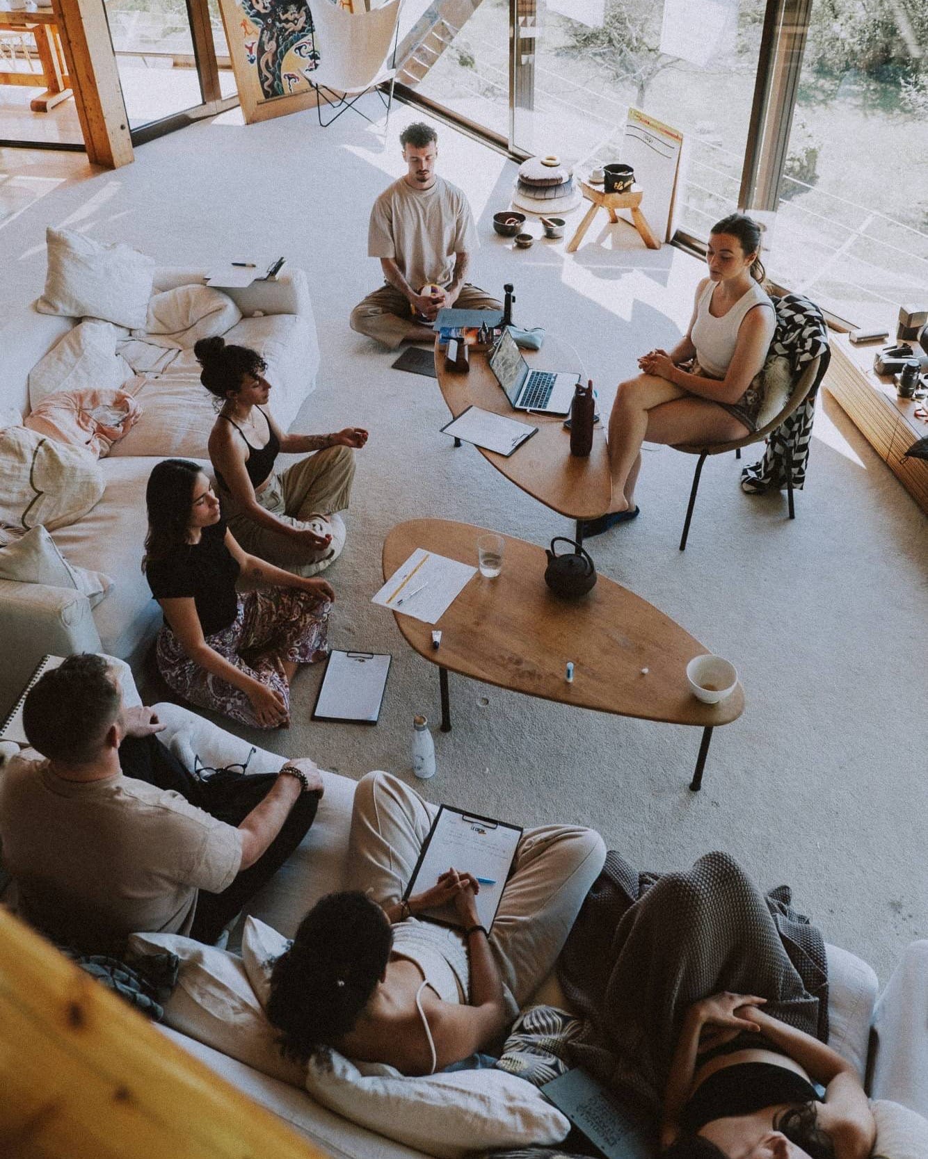 A small group sitting in meditation on cushions and sofas around low tables in a sunlit room