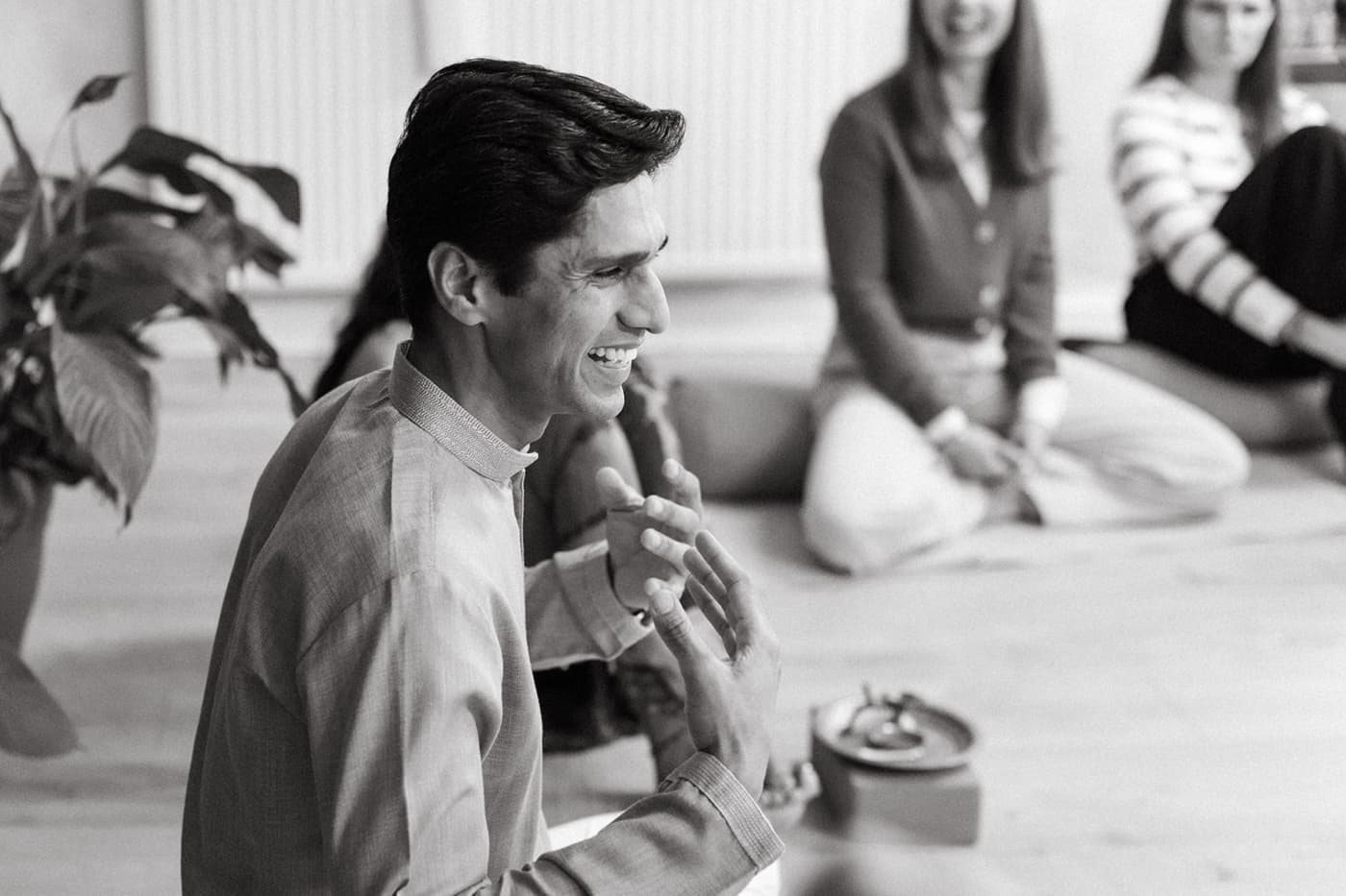 A teacher laughs and gestures while talking with students seated on cushions on a wooden floor, in black and white